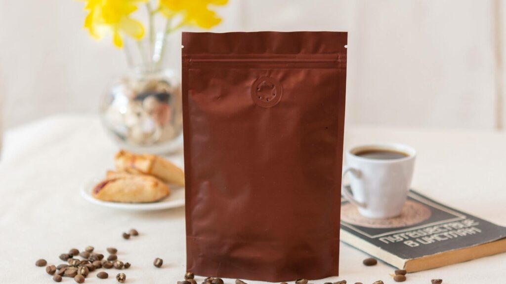 A sealed brown coffee bag with coffee beans, a cup of coffee, and pastries on a table.